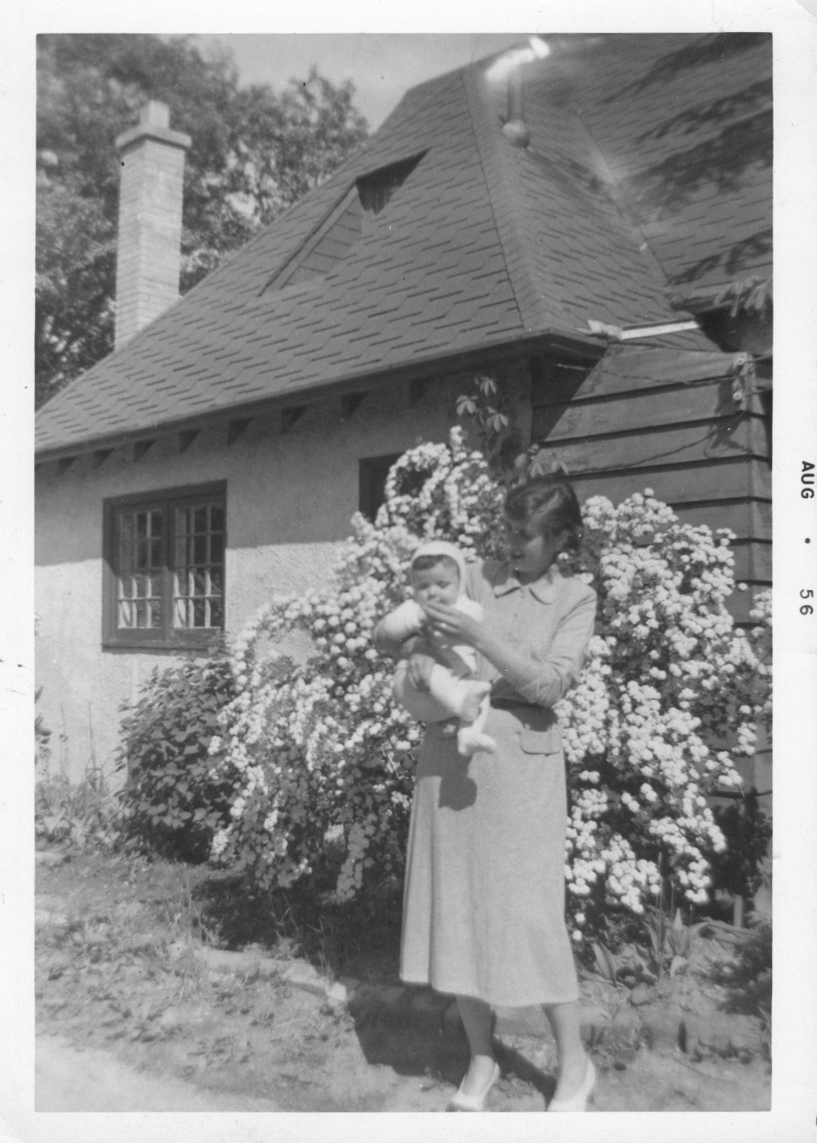 Woman holding baby outside cottage, August 1956